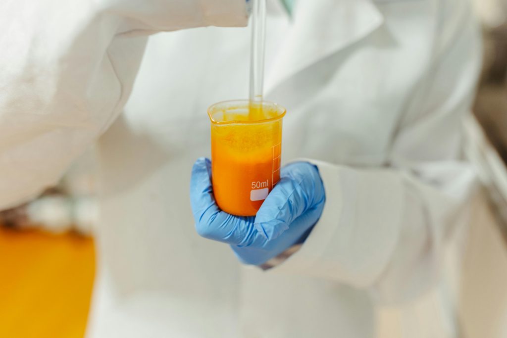 pexels photo 15831828 15831828 Close-up of a scientist in a lab coat holding a beaker with orange liquid using blue gloves.