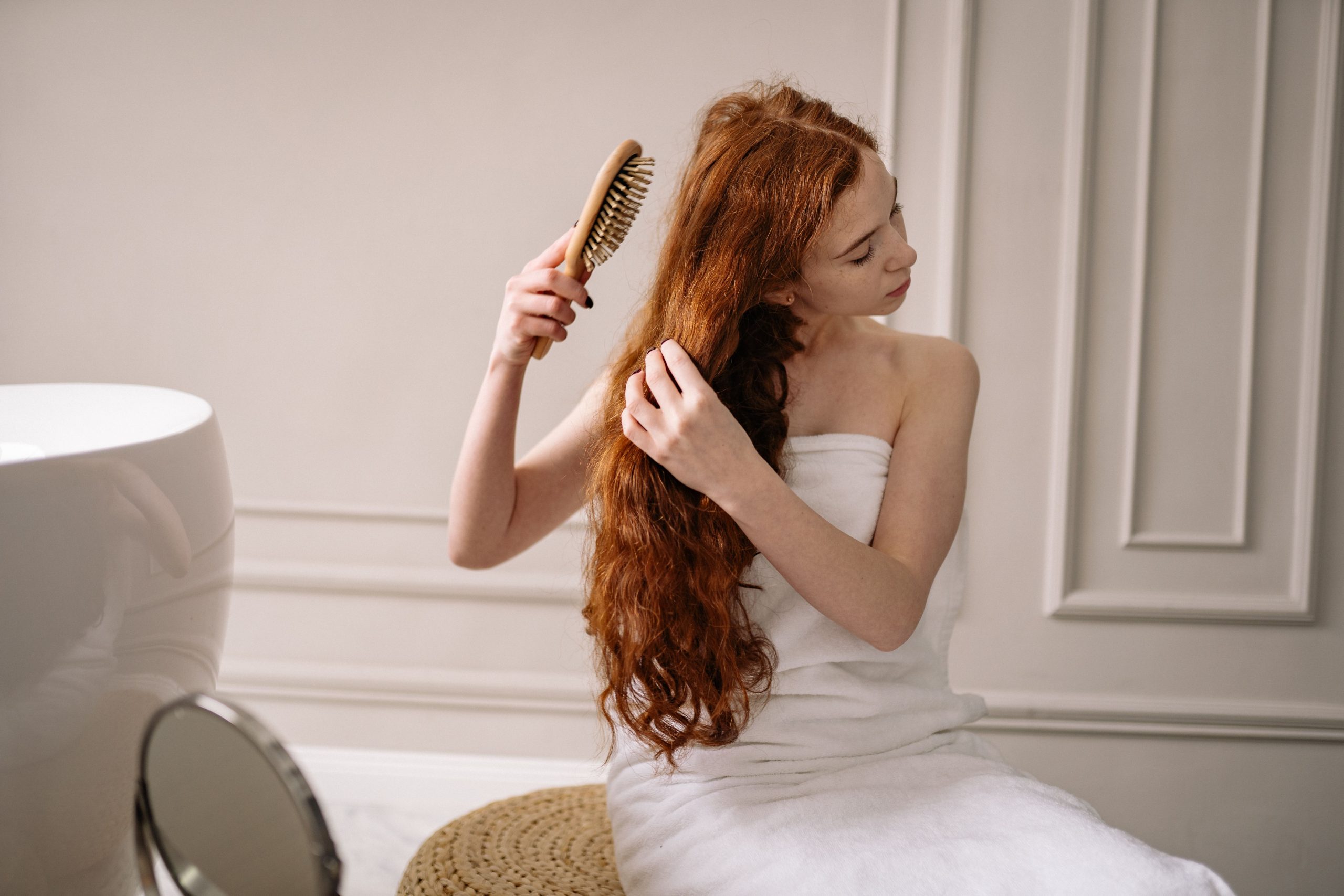 El poder de la regeneración celular: La medicina estética redefine el tratamiento contra la caída del cabello Woman with long red hair wrapped in a towel combing her hair in an elegant bathroom setting.